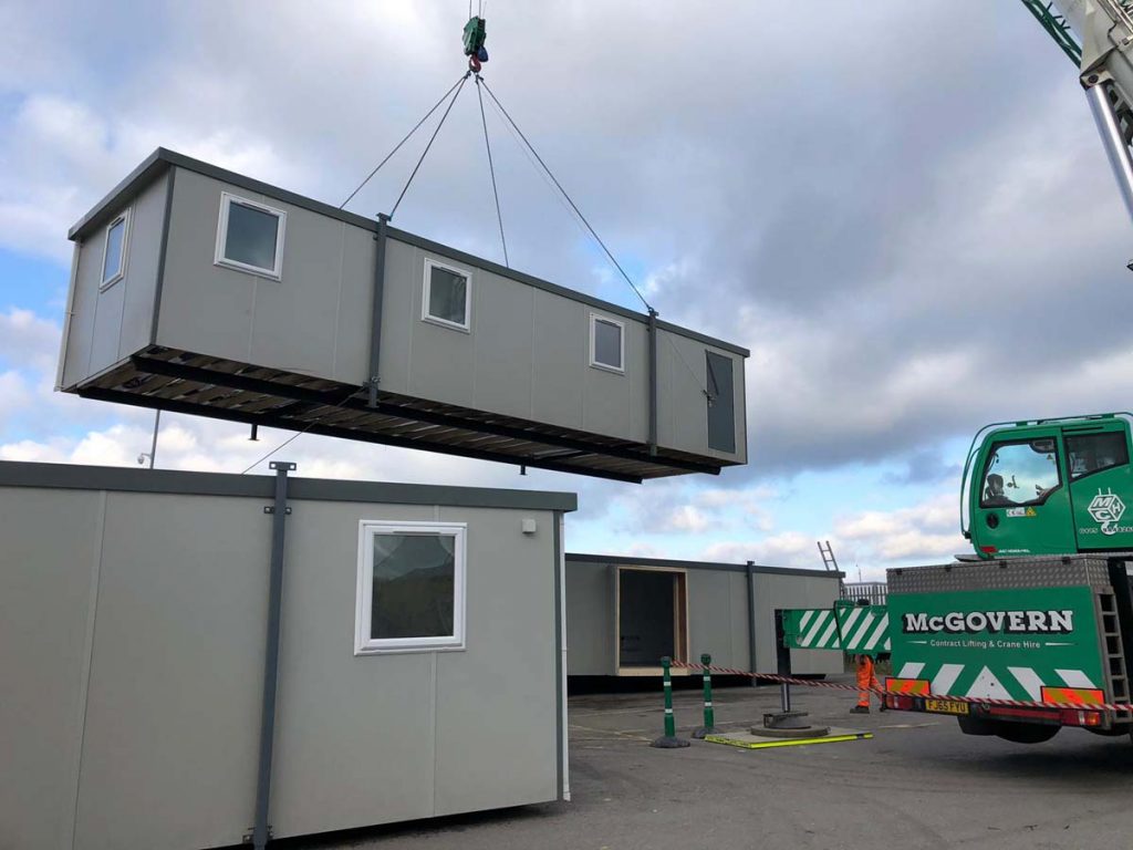 A large rectangular prefab unit is being lifted by a crane in a construction area. The crane truck, labeled "McGovern," and other prefab units are visible under a cloudy sky.