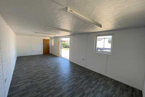 An empty room with gray wooden flooring and white walls, illuminated by ceiling lights. An open doorway and window reveal an outdoor area with grass and another building.