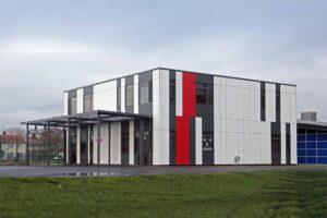 A modern, two-story building with white, gray, and red panels stands on a paved area next to a grassy lawn under a cloudy sky.