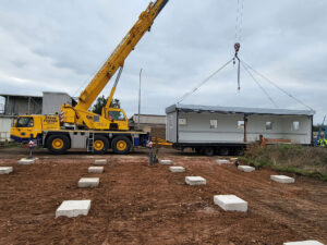 A yellow crane labeled "STEVE FOSTER CRANES" moves a white prefabricated building, dangling from cables, onto a prepared dirt construction site with concrete footings.