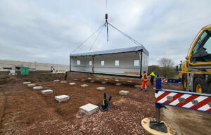 A modular building is being lifted by a crane at a construction site, with workers in safety gear guiding its placement onto prepared foundation blocks.