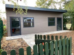 A small, modern building with two large windows and two doors, situated on a patio surrounded by a green fence, with autumn leaves scattered on the ground and trees in the background.