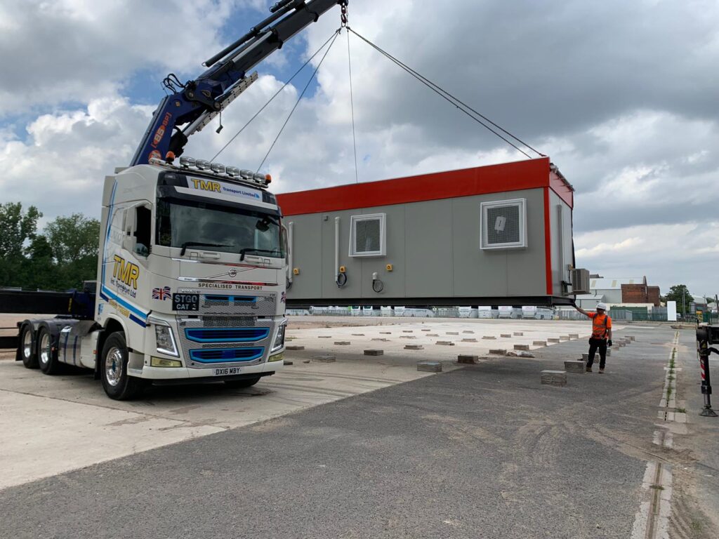 A crane truck labeled "TMR" lifts a large modular building in an industrial yard, where a worker in orange supervises the operation. Text on the truck reads "STGO CAT 3 DX16 MBY."