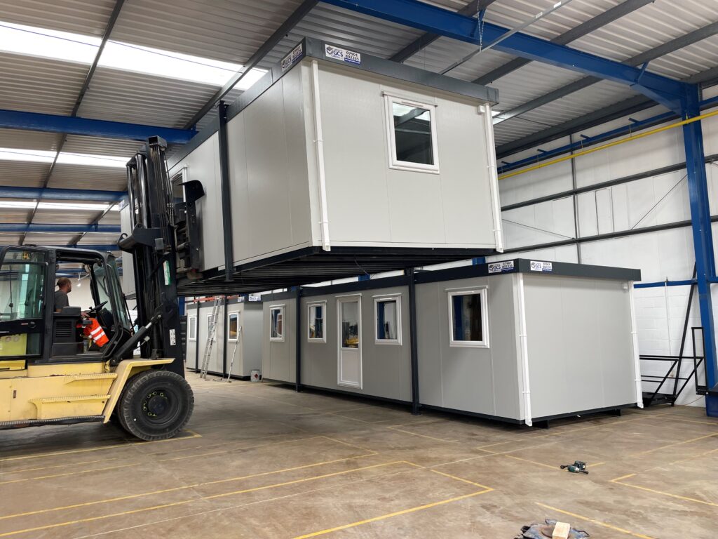 A forklift lifts a modular building unit inside a warehouse with several similar units on the ground.