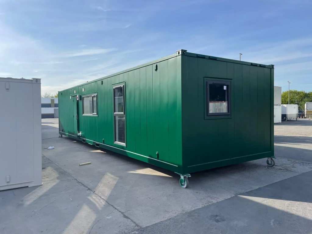 A green portable cabin with windows is parked on a concrete surface under a clear blue sky, surrounded by other similar structures.