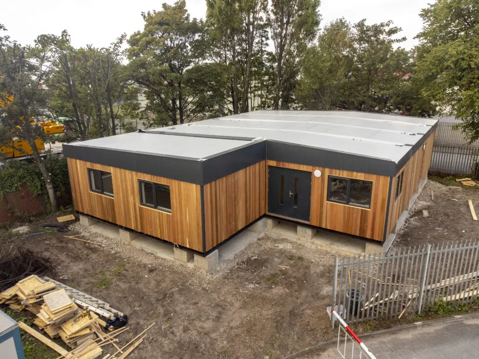 An image of a modular building with wooden panelling surrounded by trees and construction materials.