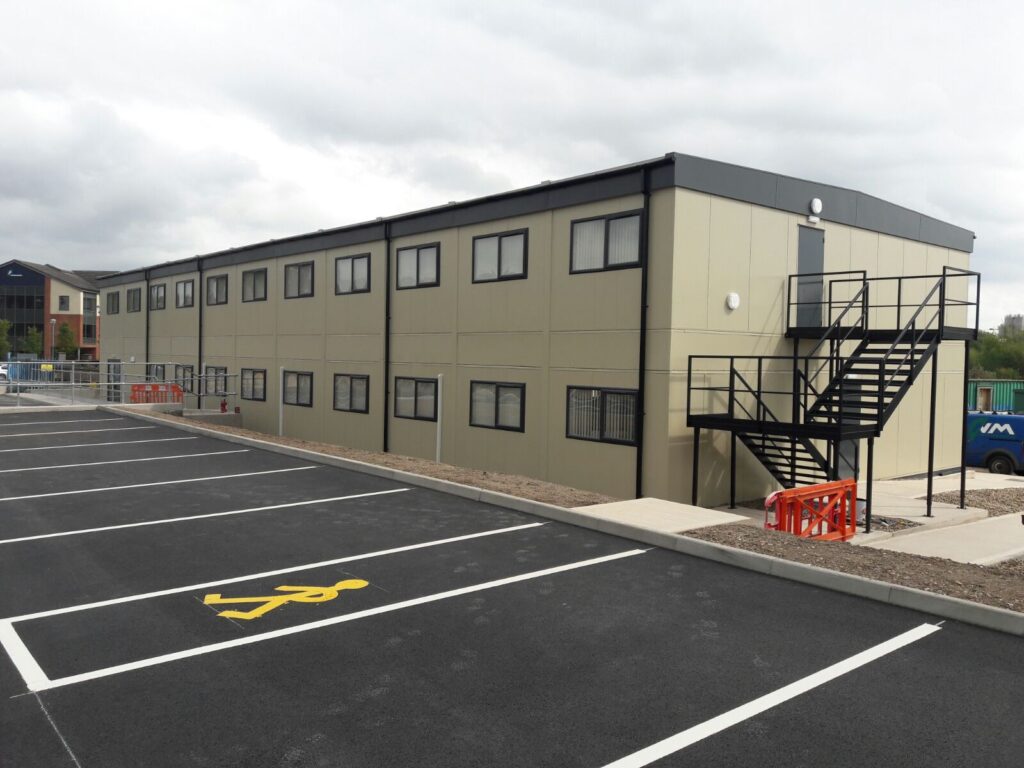 A beige, two-story portable building with a black external staircase is beside an empty, freshly painted parking lot under a cloudy sky. Orange safety barriers are near the staircase.