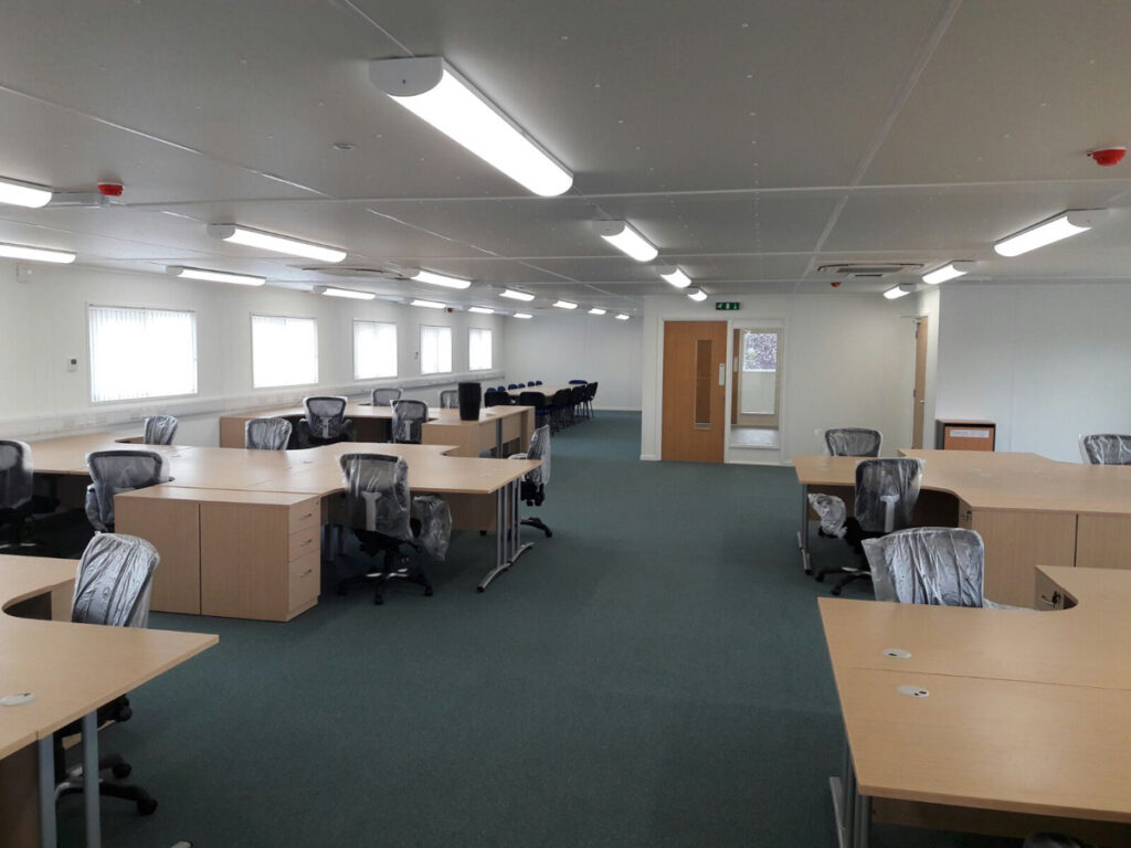 Desks and office chairs covered in plastic stand organized in rows within a well-lit, spacious office with windows lining one wall and a door at the back.