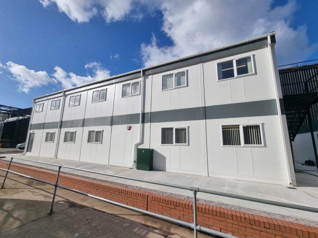 A two-story white modular building sits in an industrial area, with several windows and a red brick walkway in front. A partly cloudy sky provides the backdrop.