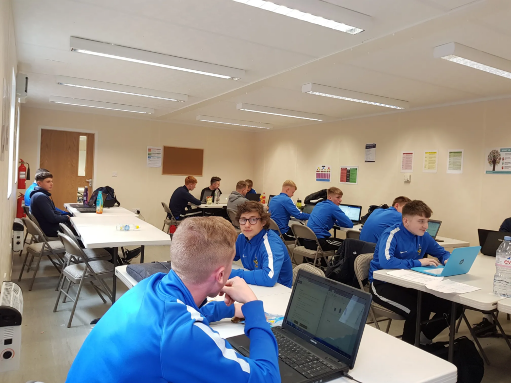 Students wearing blue tracksuits are working on laptops at tables in a classroom. The room has posters on the walls, fluorescent lights on the ceiling, and a closed door in the back.