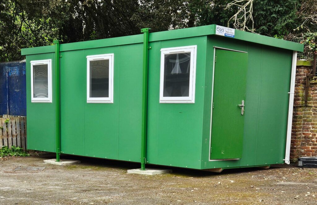 A green portable building sits on concrete blocks, featuring three white-framed windows and a door with a padlock handle. It is located outdoors, surrounded by trees and a brick wall. A sign reads "GCS 0115 7080196."