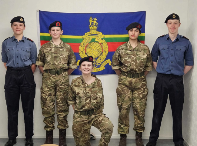 Five cadets stand and one kneels, dressed in military uniform with a Gibraltar army flag behind them, in a room with a plain white wall.