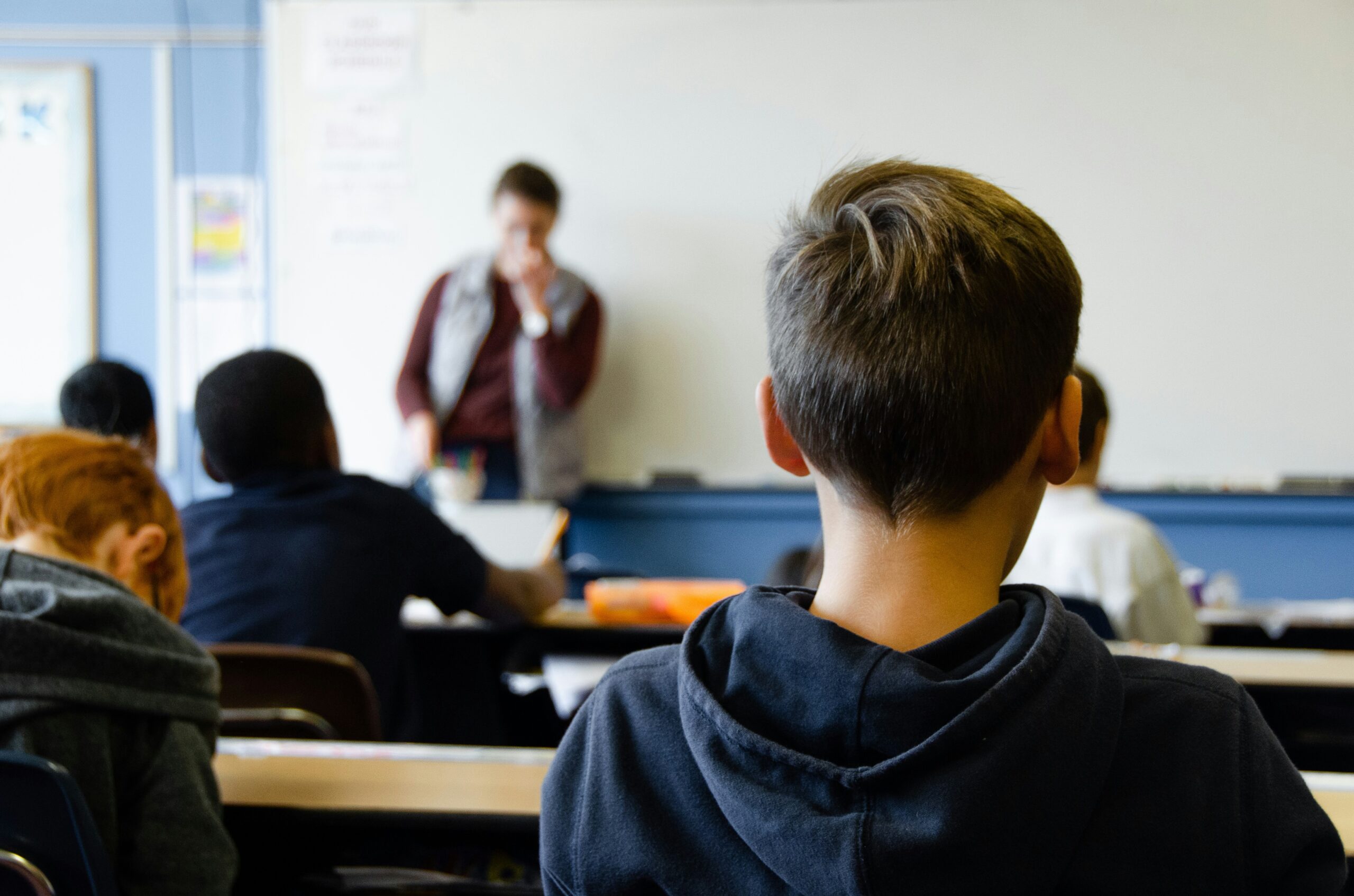 Students sit at desks facing a teacher standing at the front of a classroom, writing on a whiteboard. The classroom includes blue walls and educational posters.