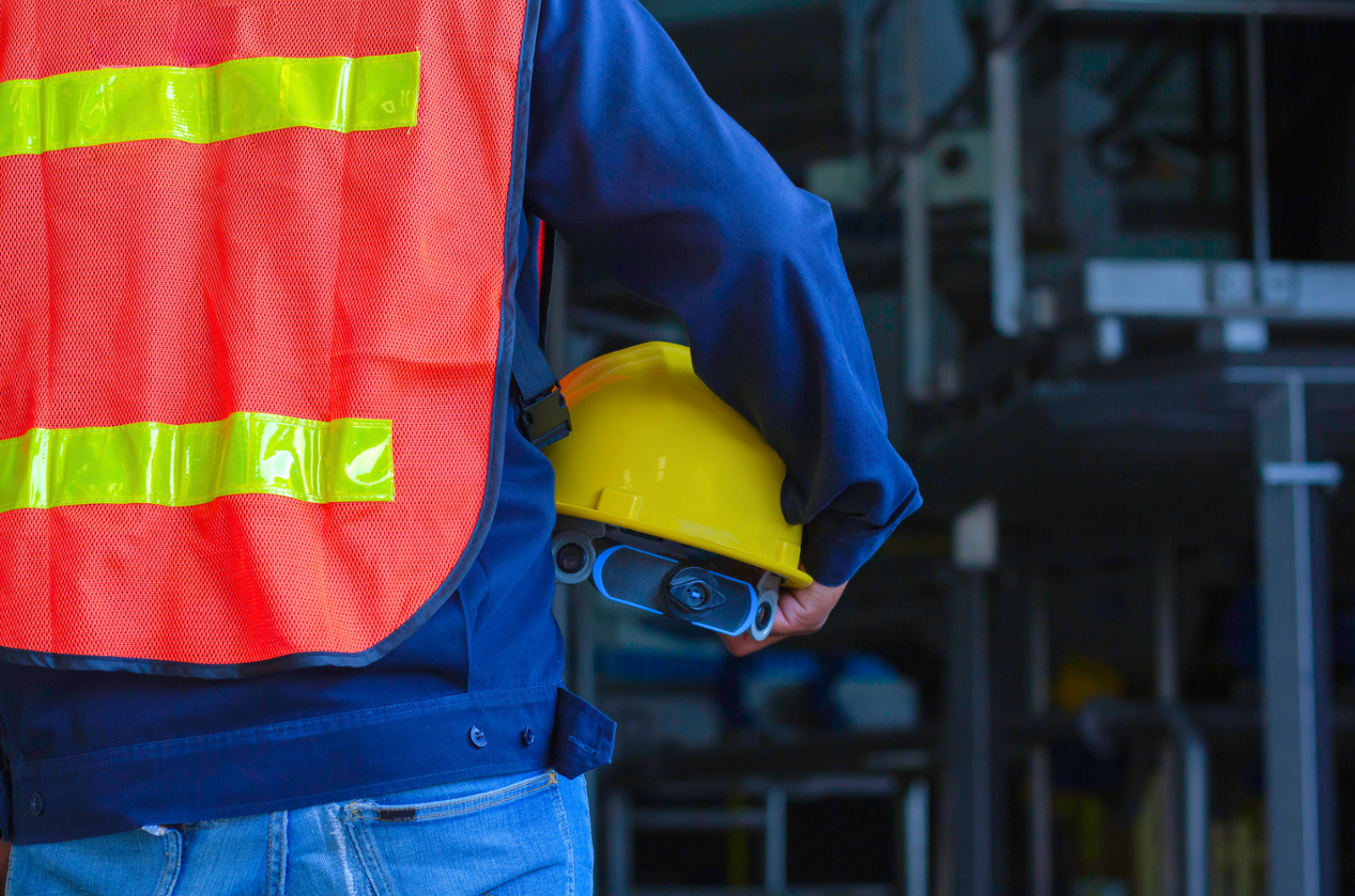 A construction worker wearing an orange safety vest and holding a hard hat under their arm.