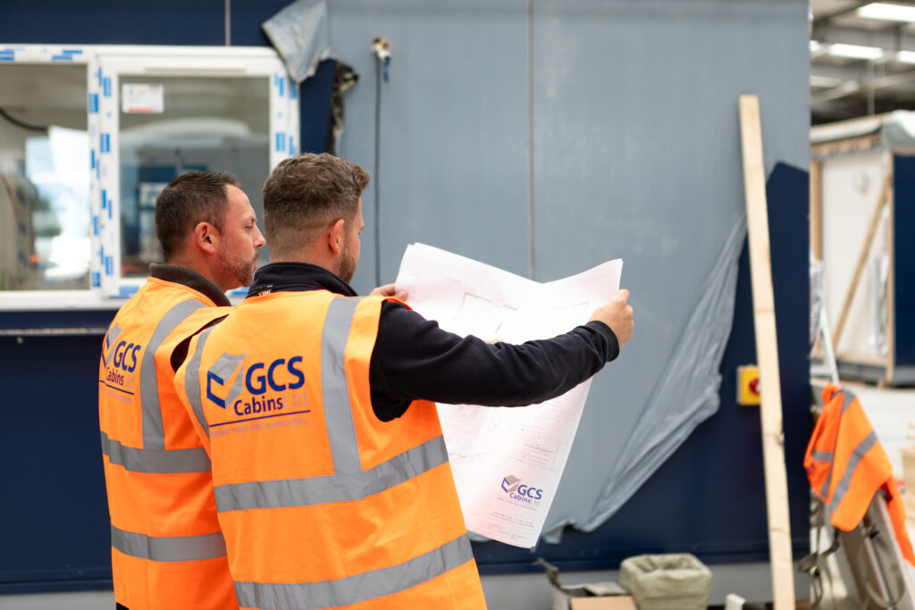 Two men in high visibility vest look at plans in a modular construction factory