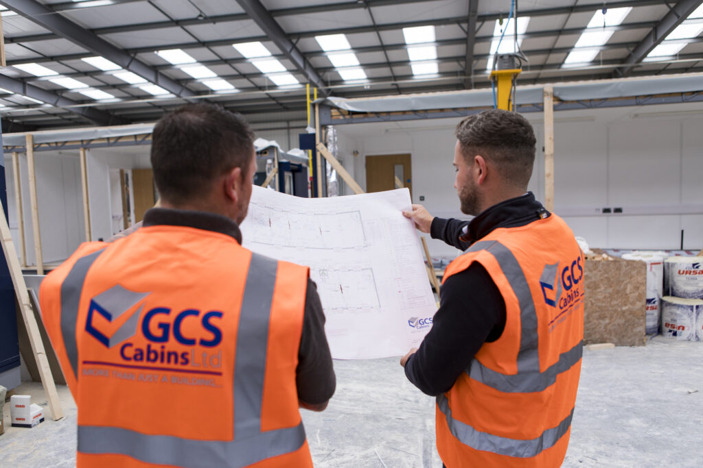 Image shows the backs of two men wearing high visibility vests looking at modular building plans in a factory